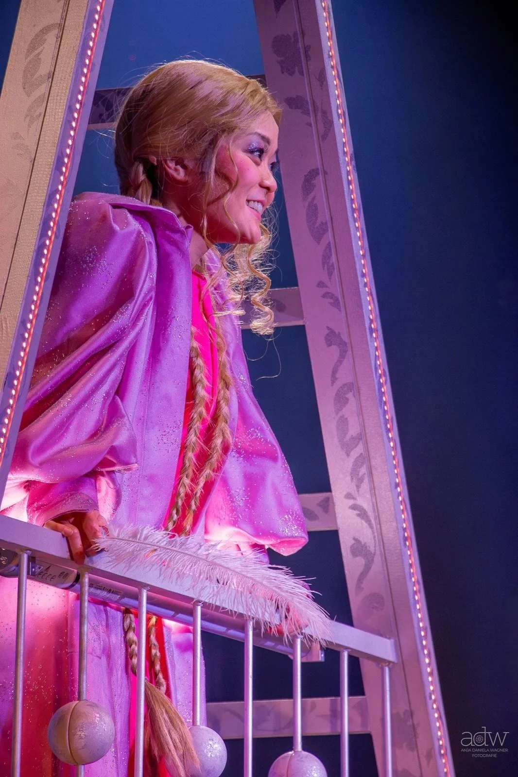 A girl dressed as Rapunzel from Disney's Tangled standing behind a pink railing, smiling, with long blonde hair in braids, surrounded by purple and pink decor.