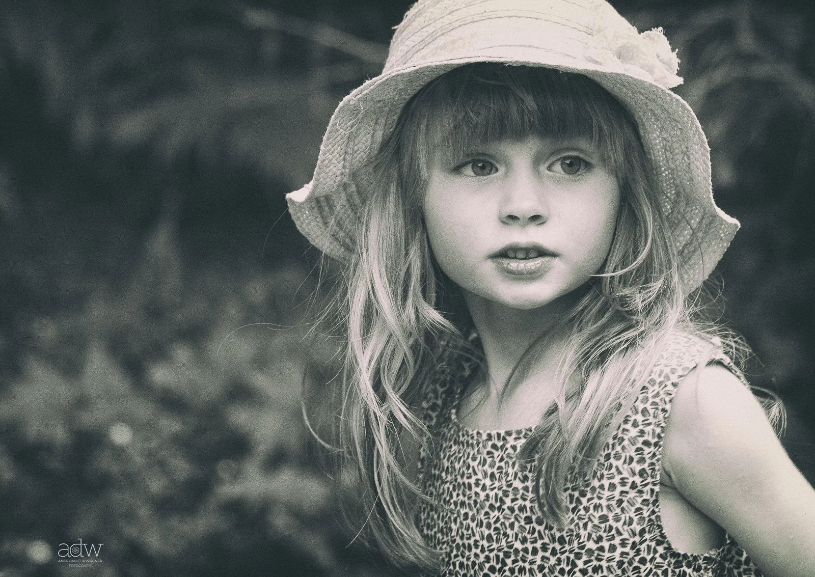 A young girl with long wavy hair wearing a sun hat and a leopard print dress, outdoors in black and white.