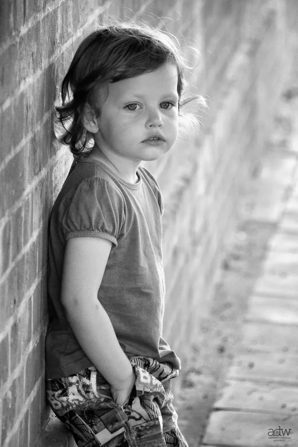 A young girl with short hair wearing a T-shirt and patterned pants, leaning against a brick wall and looking pensively at the camera.