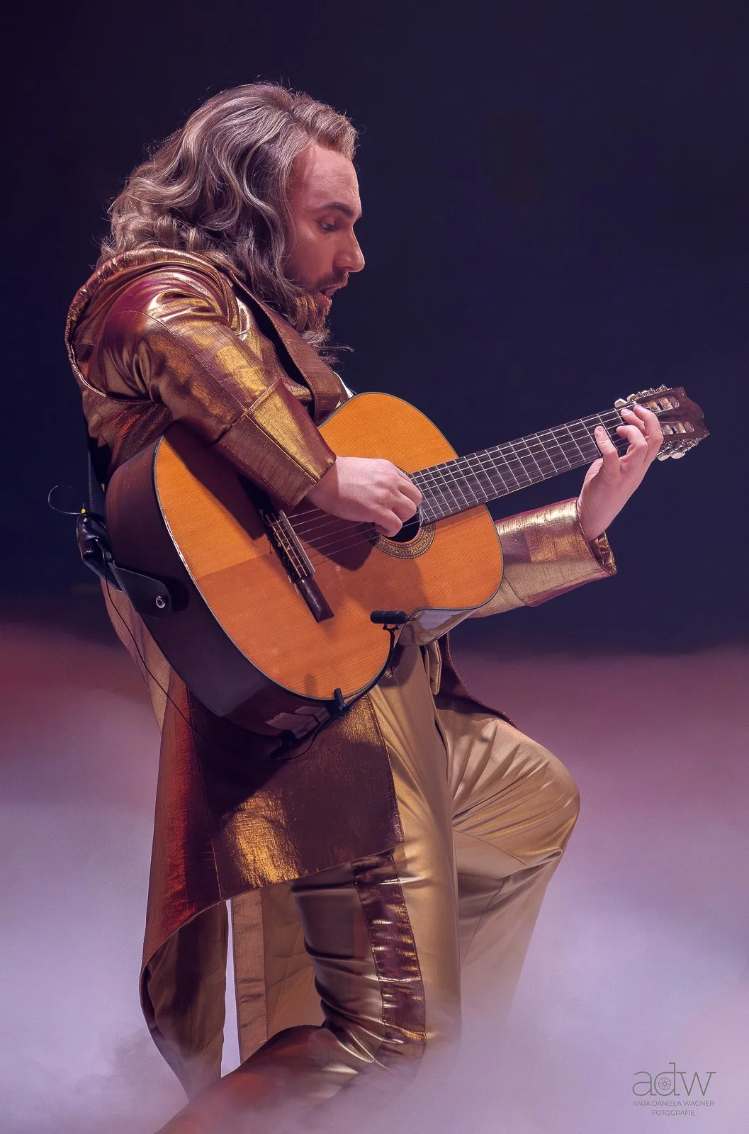 A man with long, wavy gray hair playing an acoustic guitar, dressed in a shiny gold and brown jacket and matching pants, on stage with a dark background.