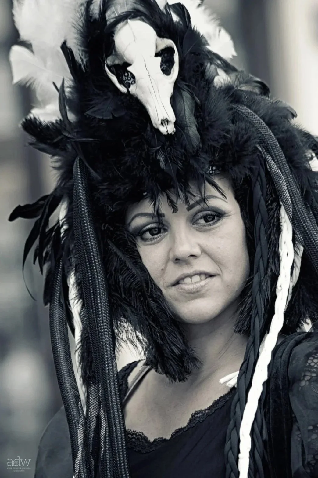 A woman in a headdress made of black feathers, braided cords, and a small animal skull, with makeup and a black lace top, looking at the camera.
