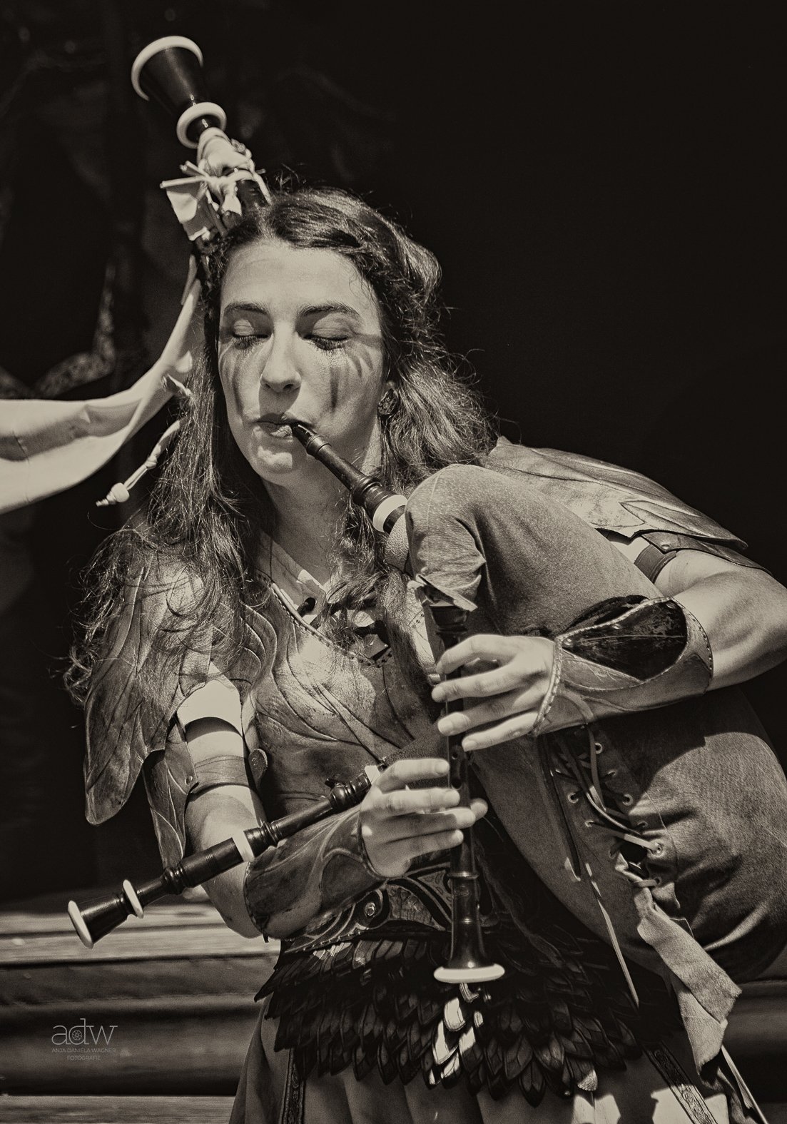 A woman with long hair playing the bagpipes on stage, dressed in costume with feathers and leather details.