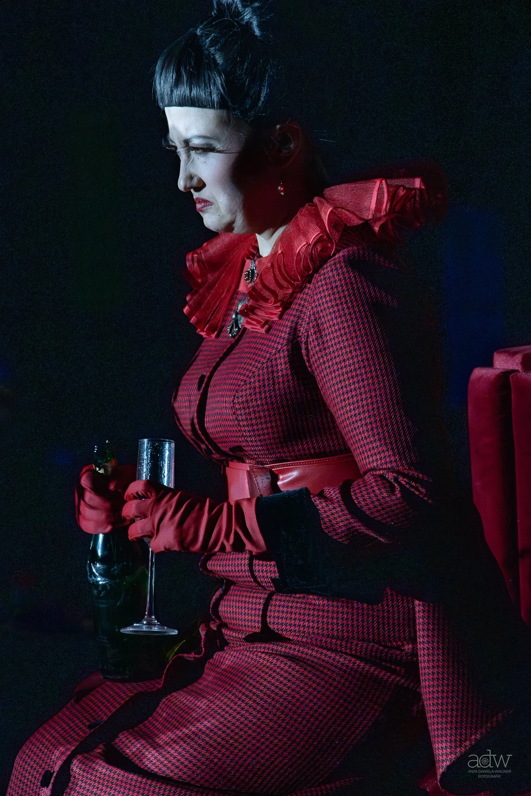 A woman with dark hair in a bob style, dressed in a red and black checkered outfit with ruffled red collar, sitting against a dark background, holding a glass of champagne and a bottle, wearing long red gloves and earrings.