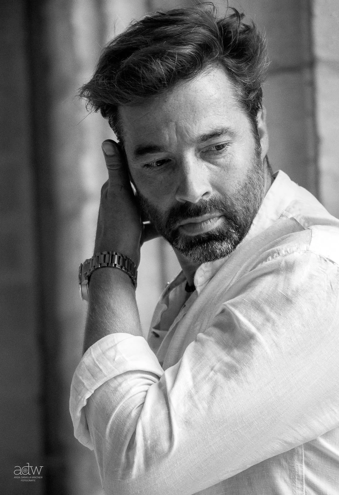 A black and white photo of a man with a beard and styled hair, wearing a light-colored shirt, looking down and touching the back of his head with his left hand, which has a watch on the wrist.