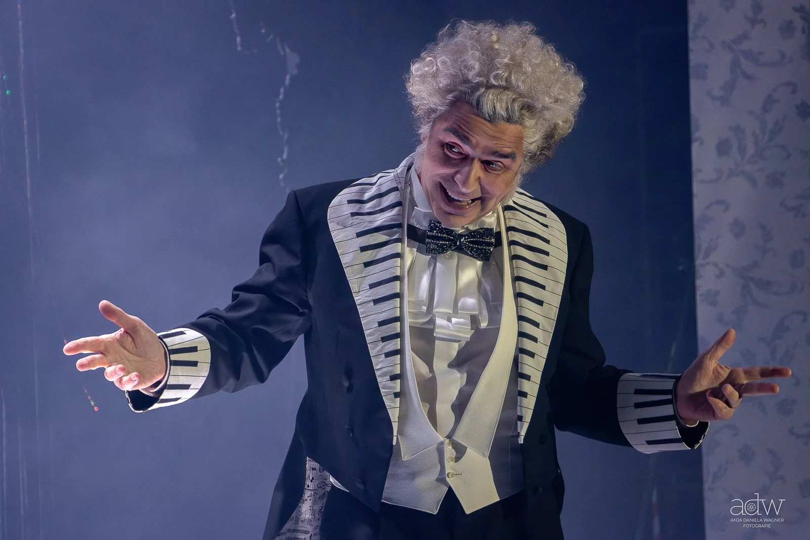 An elderly man dressed as a clown or mime, wearing a black tuxedo with striped lapels and cuffs, a white ruffled shirt, and a glittery bow tie, making expressive gestures with his hands in front of a dark background.