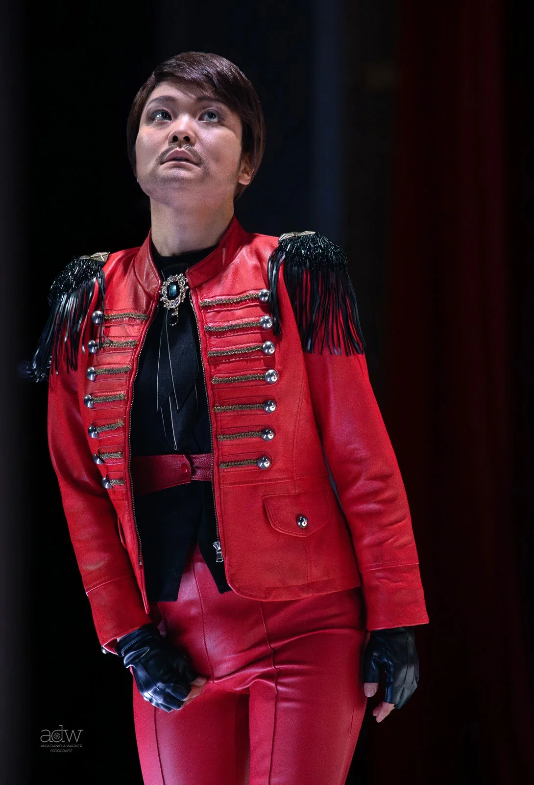 Person wearing a red military-style jacket with black fringe epaulets, decorative metallic buttons, and matching red pants, standing against a dark background.