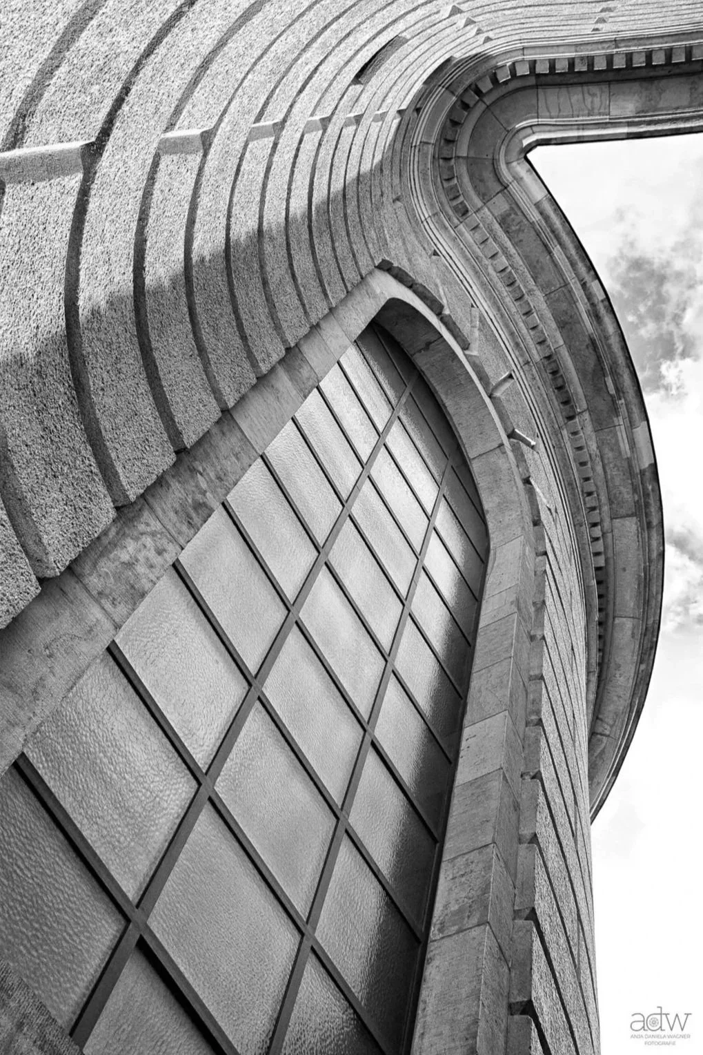 Black and white photograph of a curved building facade, showing large rectangular windows and stone architectural details. The perspective is from below, looking up at the building.
