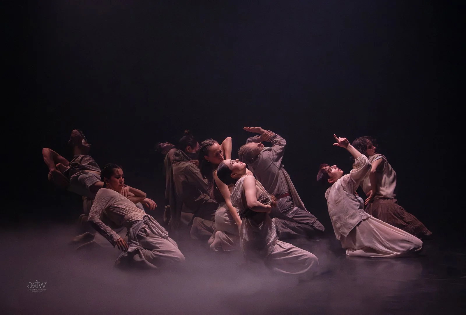 A group of dancers in neutral-toned costumes performing on stage with a dark background and fog, in various contemplative poses.