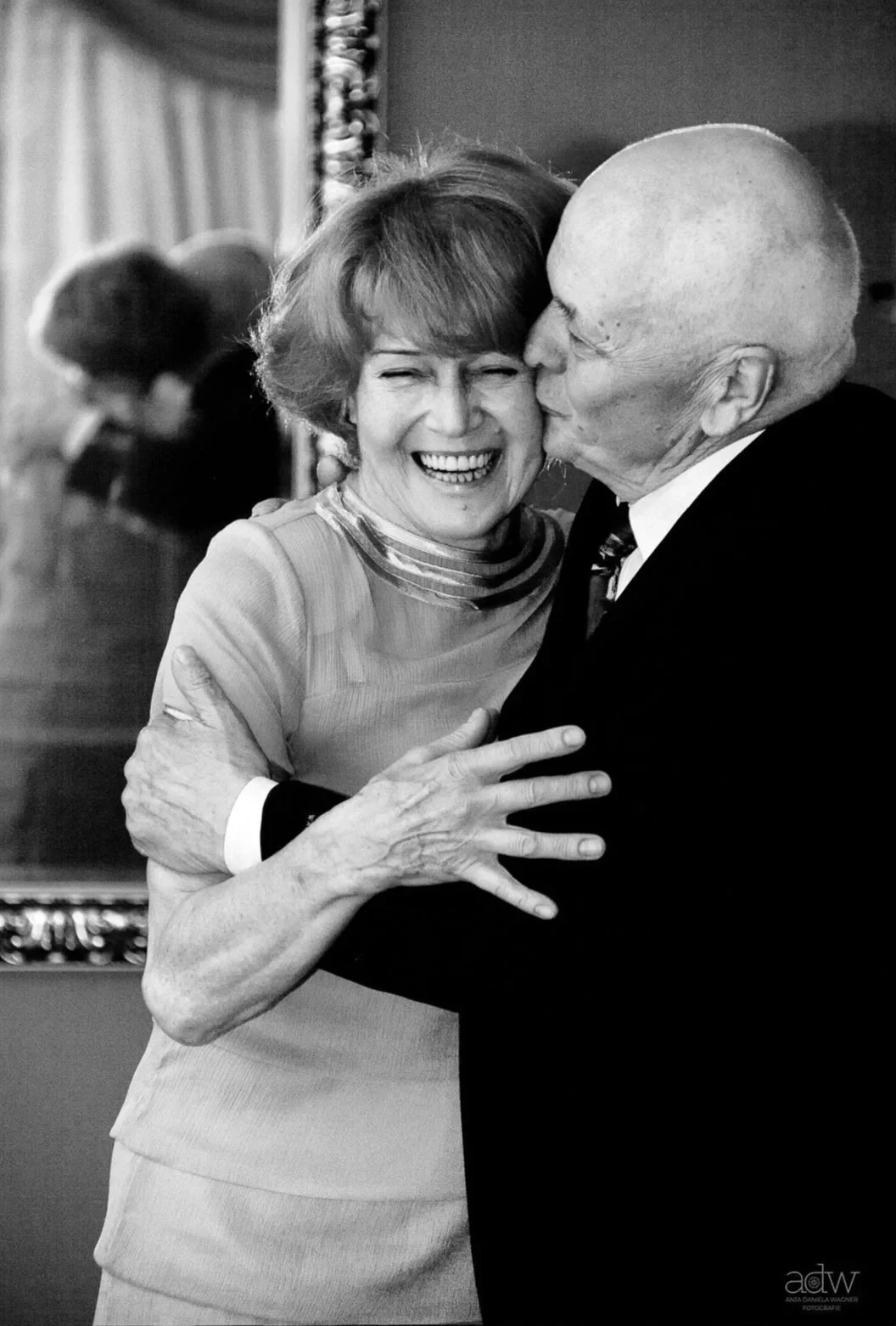 A black and white photo of a woman and a man sharing a joyful embrace, with the man kissing the woman's cheek, both smiling widely. The woman has short hair and is wearing a dress with a high neck, while the man is bald and wearing a suit and tie.