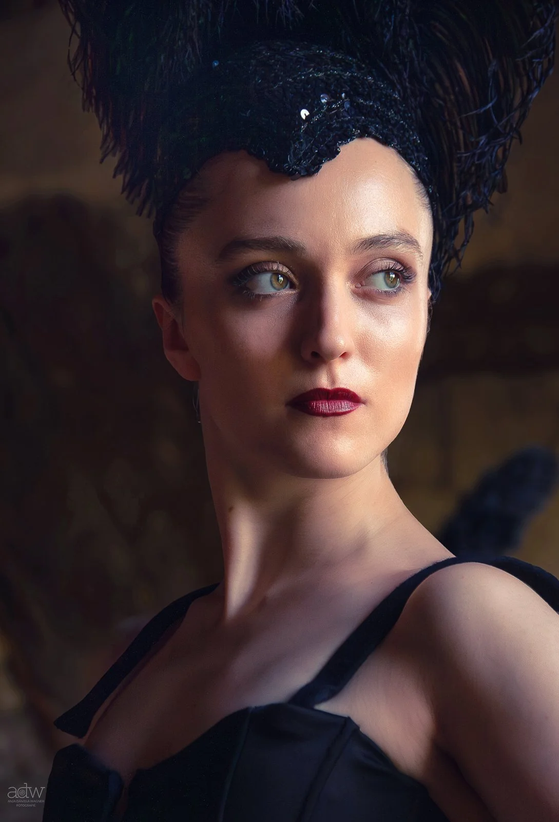 A woman with light skin, blonde hair, piercing green eyes, dark makeup, and red lipstick wearing a dramatic black headdress with feathers and a black dress with straps.