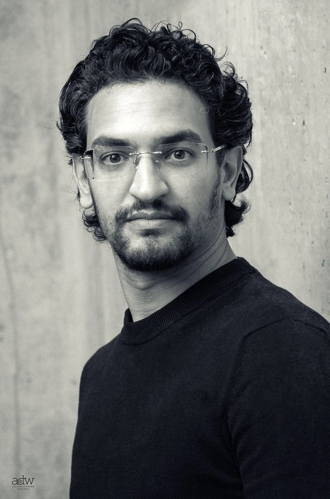 Black and white portrait of a man with curly hair, glasses, a beard, and mustache, wearing a black shirt, standing against a plain wall.