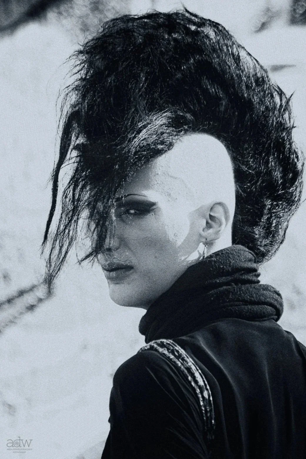 A woman with a mohawk hairstyle, dark makeup, earrings, and wearing a jacket and hoodie, looking over her shoulder in a black-and-white photo.