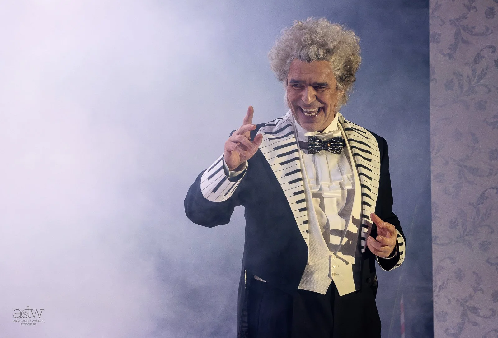 An elderly man with curly gray hair wearing a tuxedo with a piano keyboard pattern on the lapel and sleeves, a white ruffled shirt, and a glittery black bow tie, smiling and gesturing with his fingers.