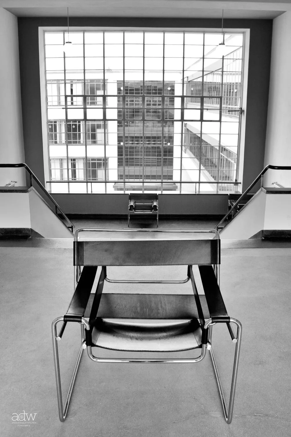 Black and white photo of a modern stairwell with two black chairs, large grid-patterned window, and building exterior visible outside.