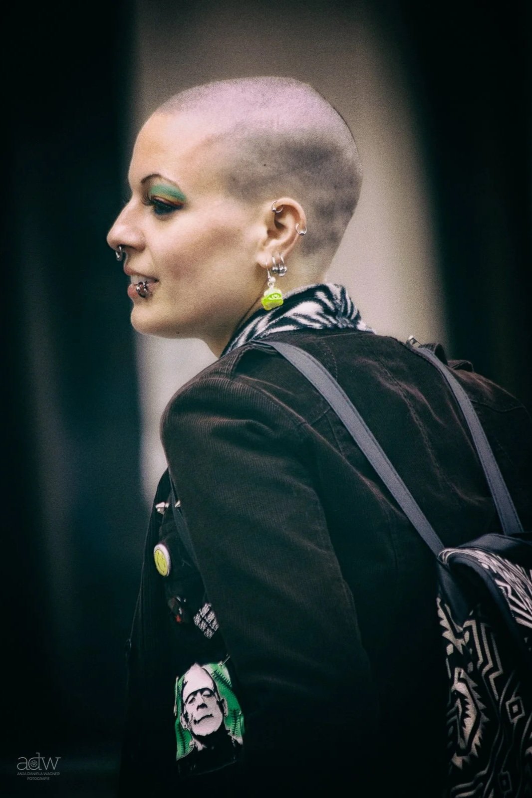 A person with a shaved head and multiple earrings, wearing makeup with green eyeshadow, and a jacket with cartoon patches, carrying a backpack.