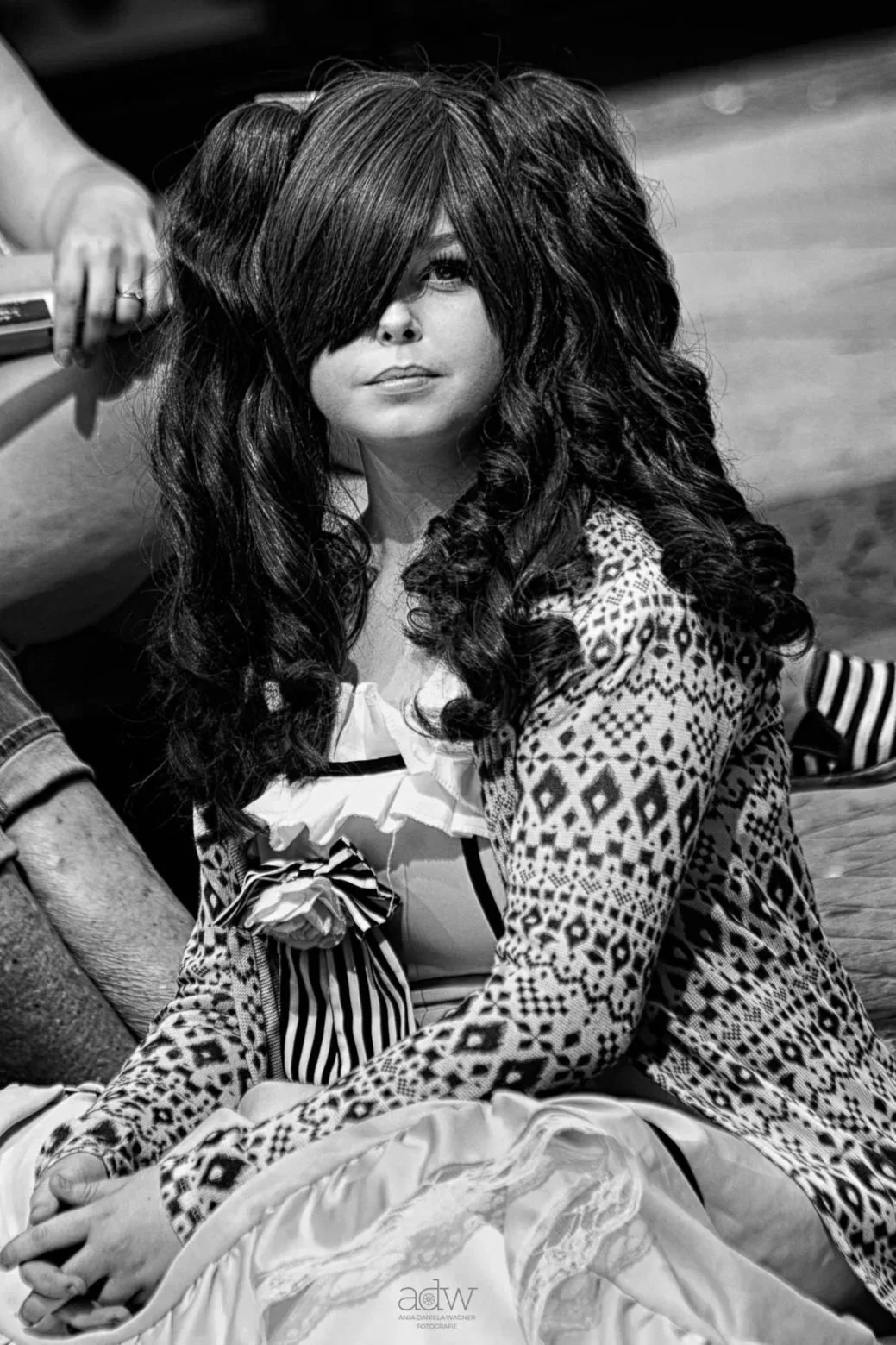 A woman with long, curly hair and bangs, wearing patterned clothing, sitting indoors with a serious expression.