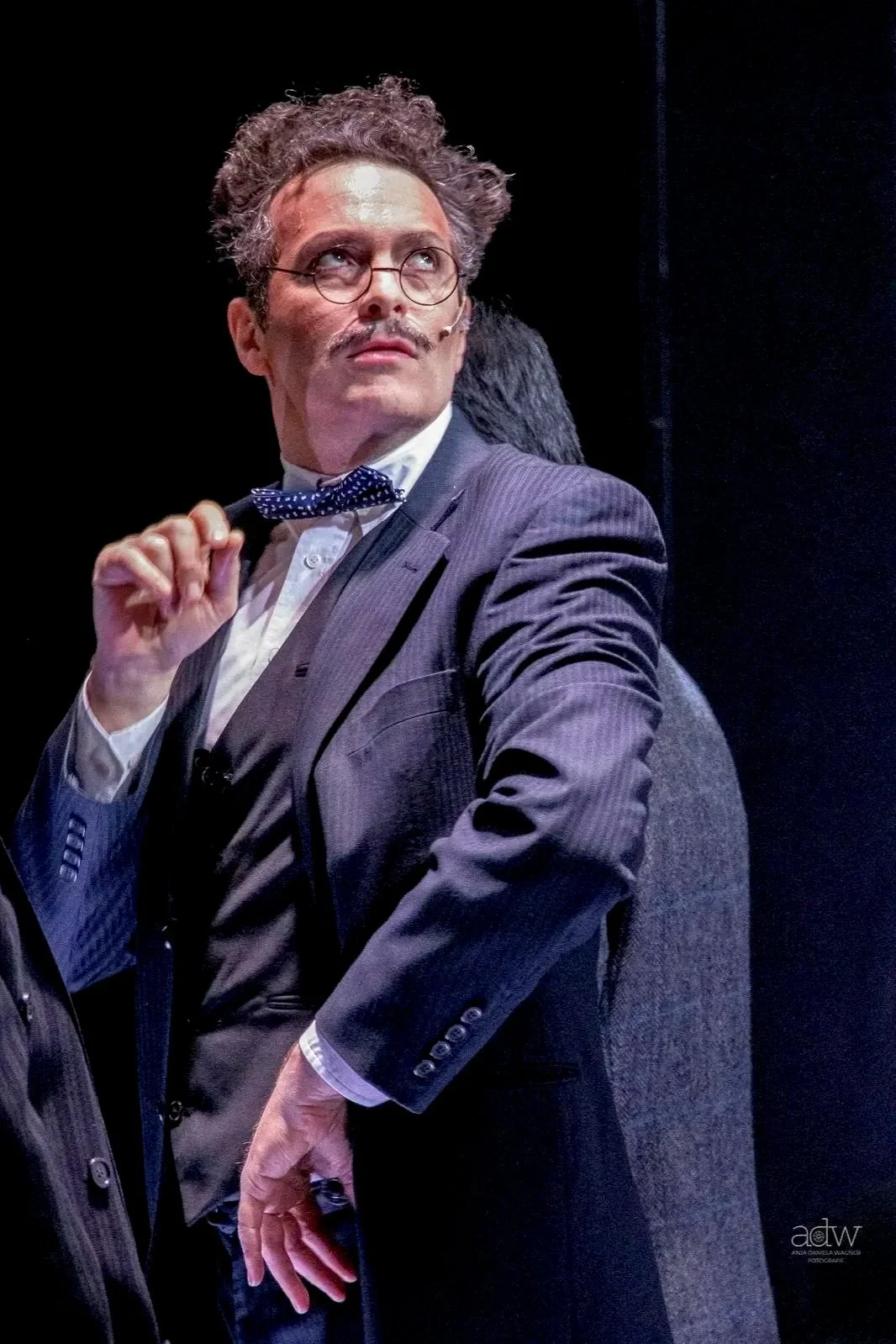 A man dressed in a gray suit, wearing glasses, with a bow tie, sits on a stage with a black background, and appears to be speaking or performing.