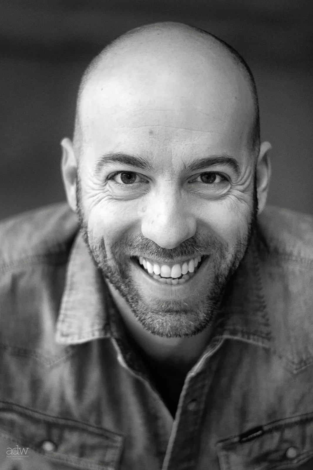 A black and white close-up portrait of a smiling man with a bald head and facial hair, wearing a denim shirt.