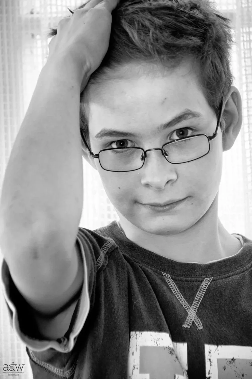 Young person with short hair and glasses, looking directly at the camera, with one hand resting on their head. They are wearing a dark t-shirt.