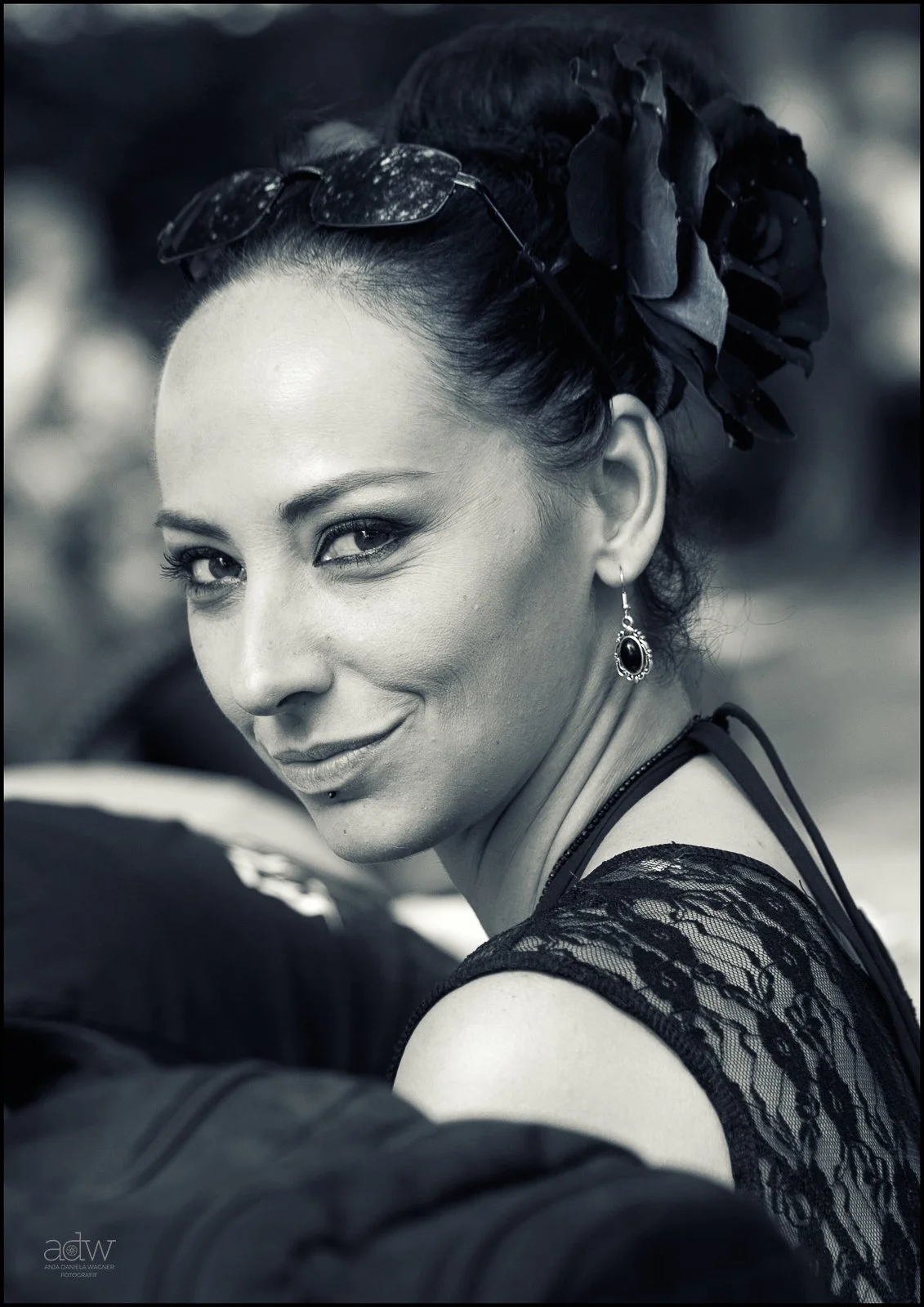 A woman with dark hair styled in a bun adorned with a large black flower, wearing sunglasses on her head, lace top, and dangling earrings, smiling and looking over her shoulder.