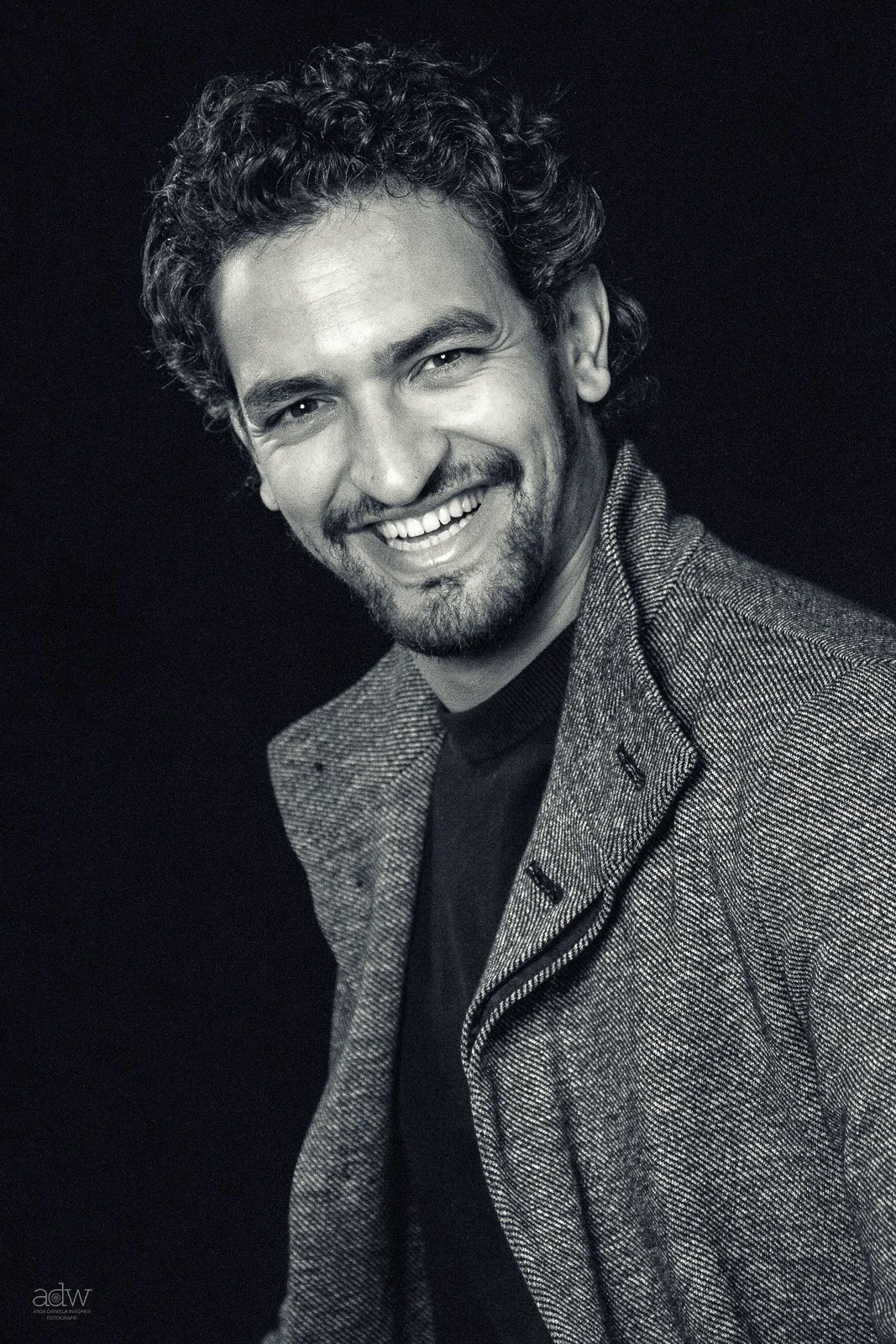 A black-and-white photo of a man with curly hair and a beard smiling, dressed in a checkered blazer over a dark shirt.