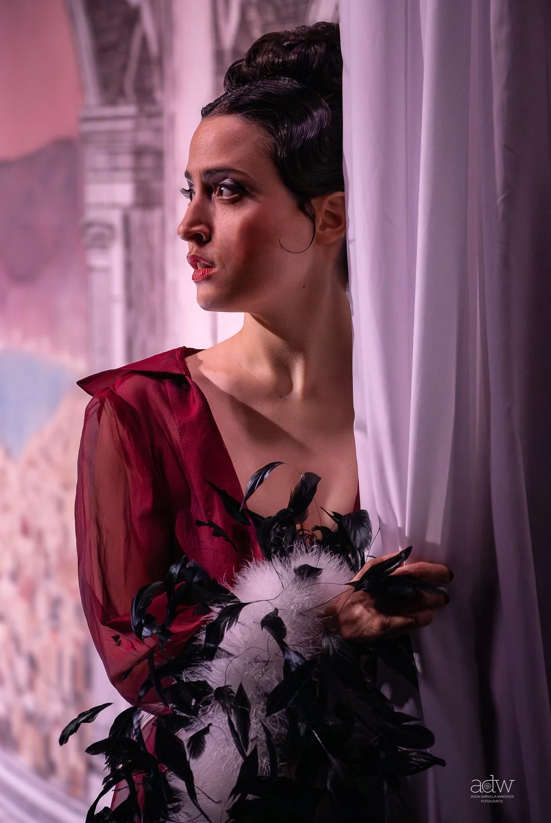 A woman with dark hair styled up, wearing a deep red sheer top with black feathered accessories, standing near a white curtain and gazing outside with a thoughtful expression.