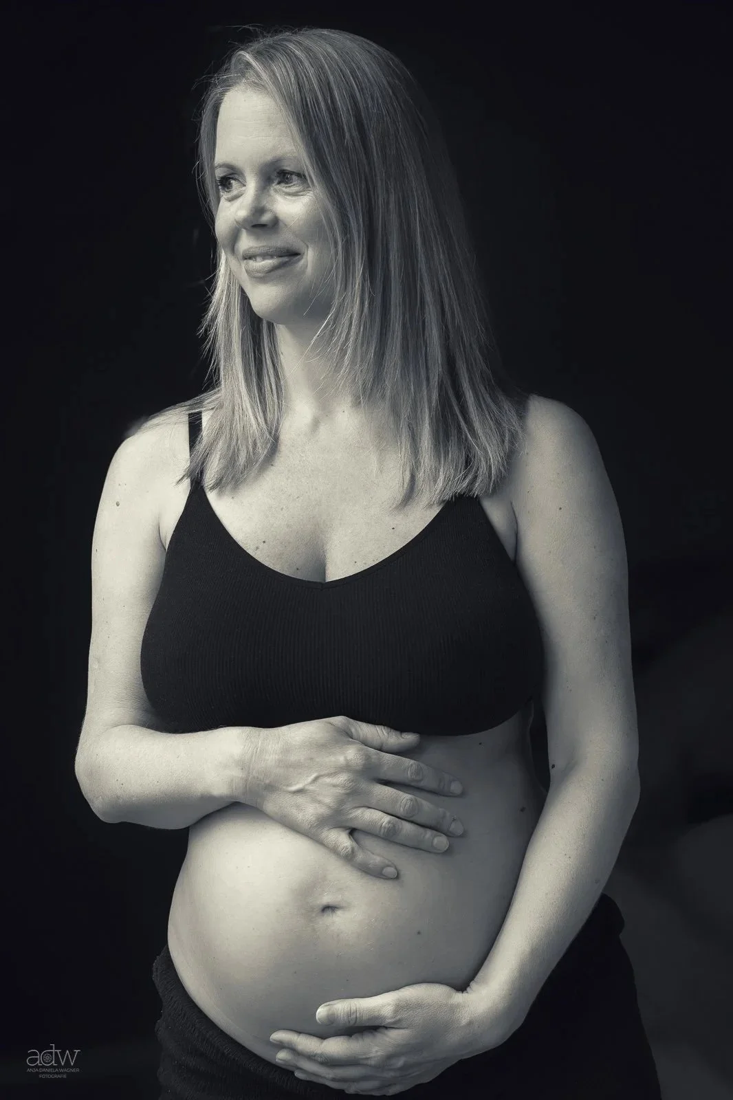 Black and white portrait of a pregnant woman with shoulder-length hair, wearing a black tank top, smiling softly, with her hands gently resting on her belly.