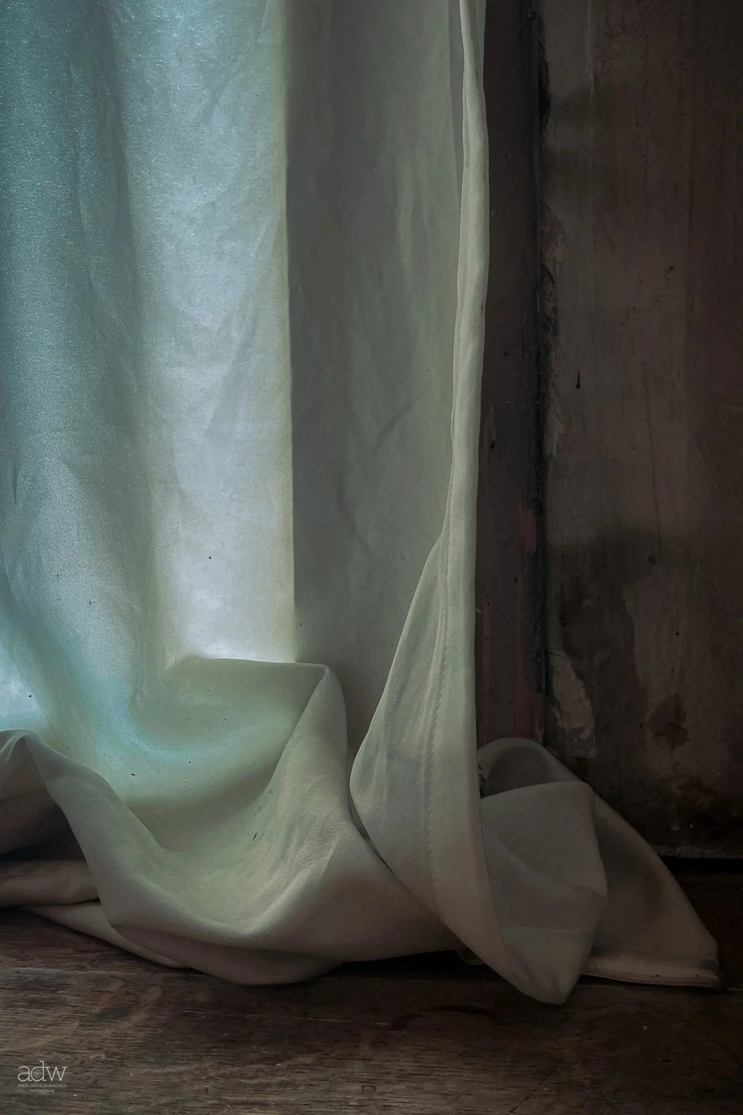 Close-up of a cream-colored curtain draped on an old wooden floor next to a weathered concrete wall.