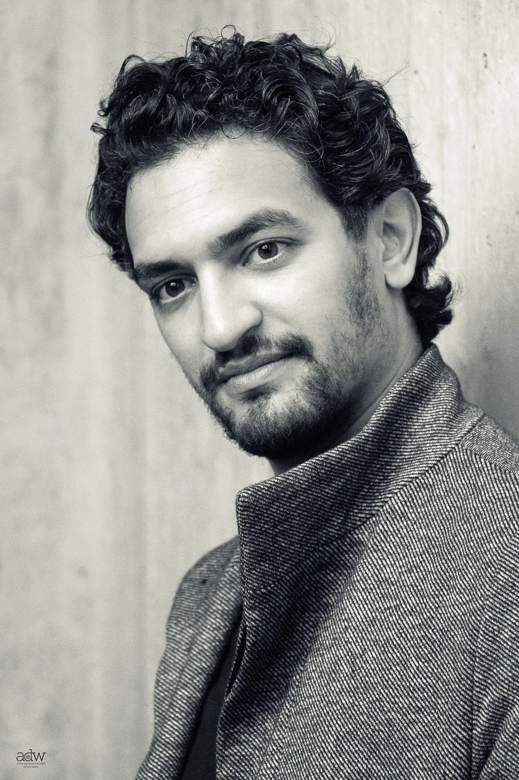 Black and white portrait of a young man with curly hair and facial hair, wearing a textured jacket, looking at the camera with a slight smile.