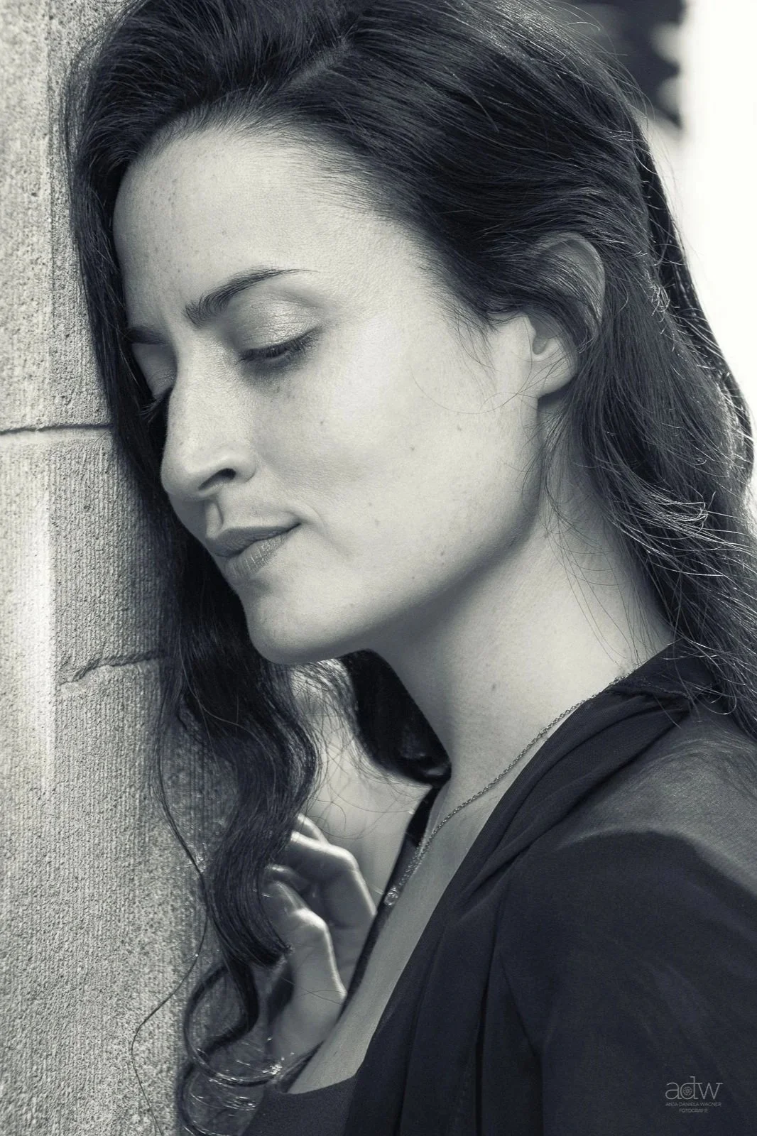 A black-and-white photo of a woman with long dark wavy hair, her eyes closed, leaning her face against a textured wall. She appears serene and contemplative.