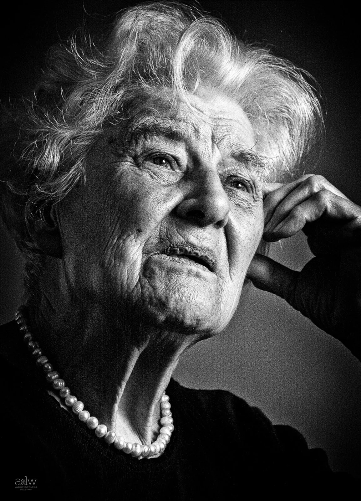 Black and white close-up portrait of an elderly woman with gray hair, wearing a pearl necklace, resting her head on her hand, looking thoughtful.