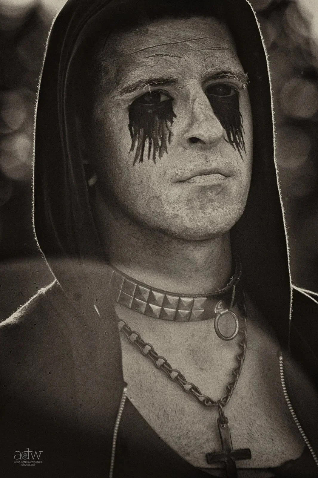 Black and white close-up of a person with dark makeup around their eyes, wearing a hood, a spiked choker, and a chain necklace with a cross pendant.