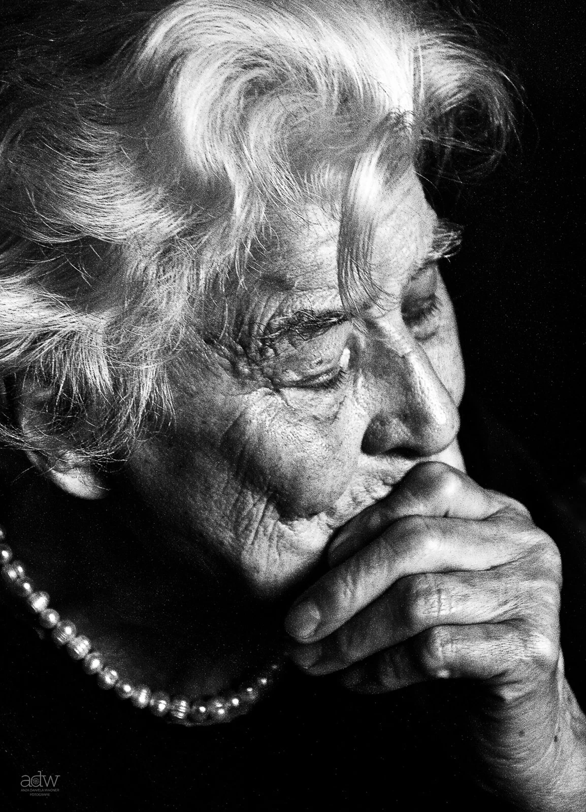 Close-up black and white photo of an elderly woman with wavy hair, wearing a pearl necklace, with her eyes closed and hand near her mouth, appearing to be deep in thought.
