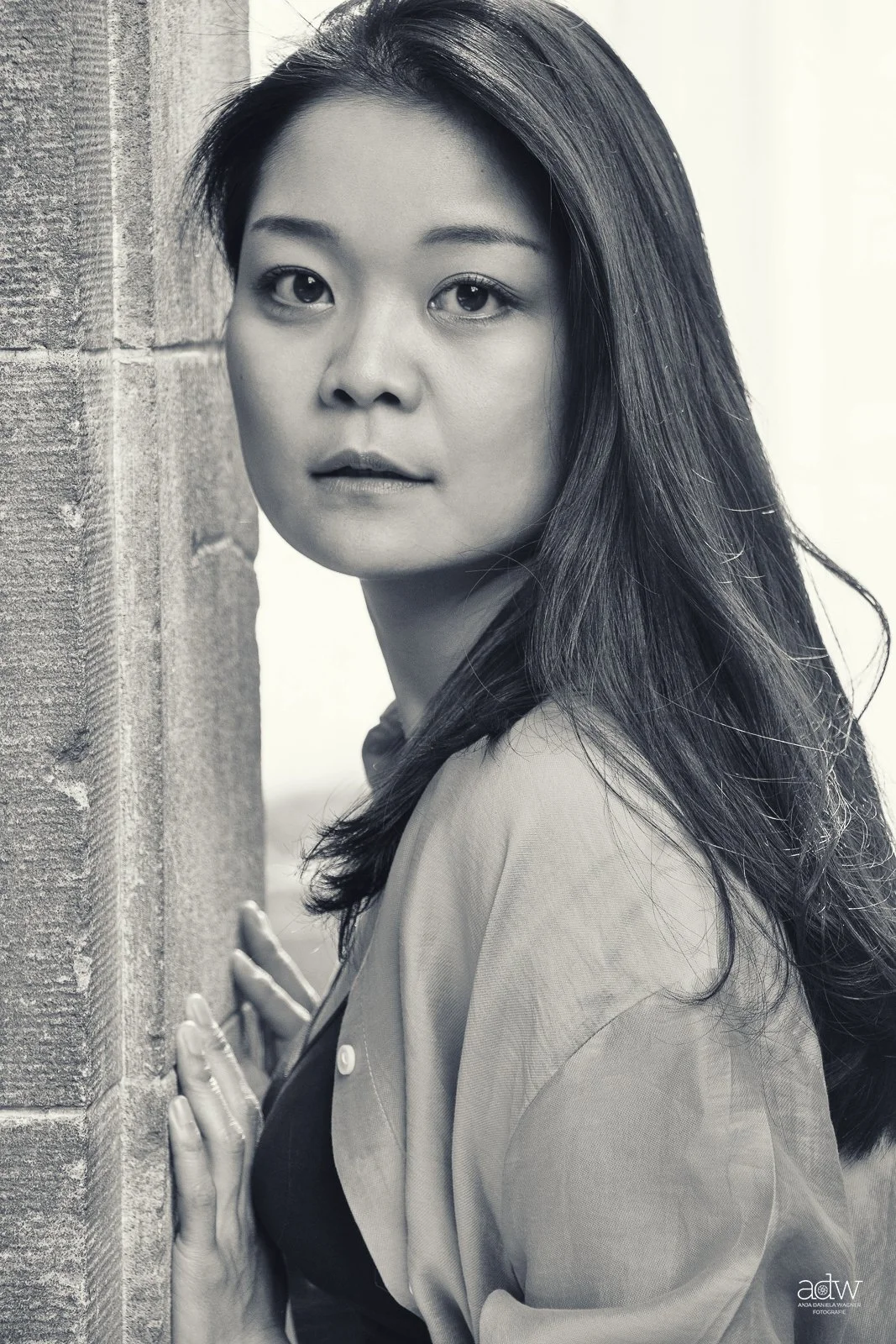 Black and white portrait of an Asian woman with long hair, leaning against a wooden post, looking at the camera.