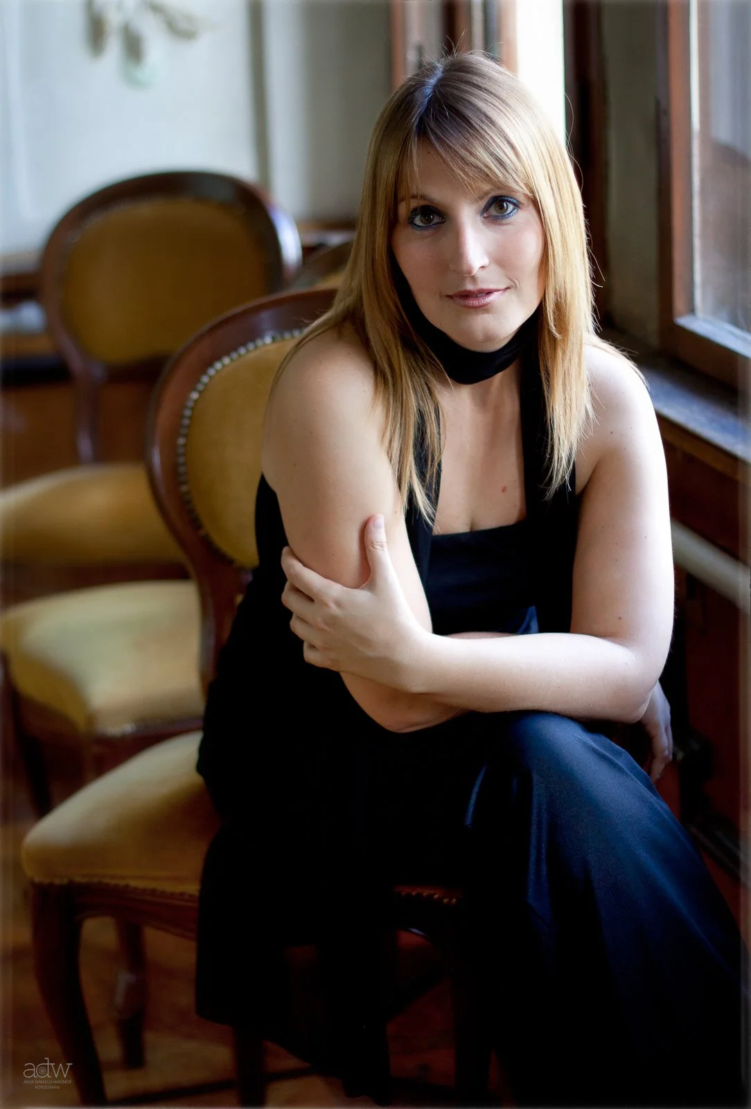 A woman with blonde hair and blue eyes sitting on a chair near a window, wearing a black sleeveless top and pants, with her arms crossed and looking at the camera.