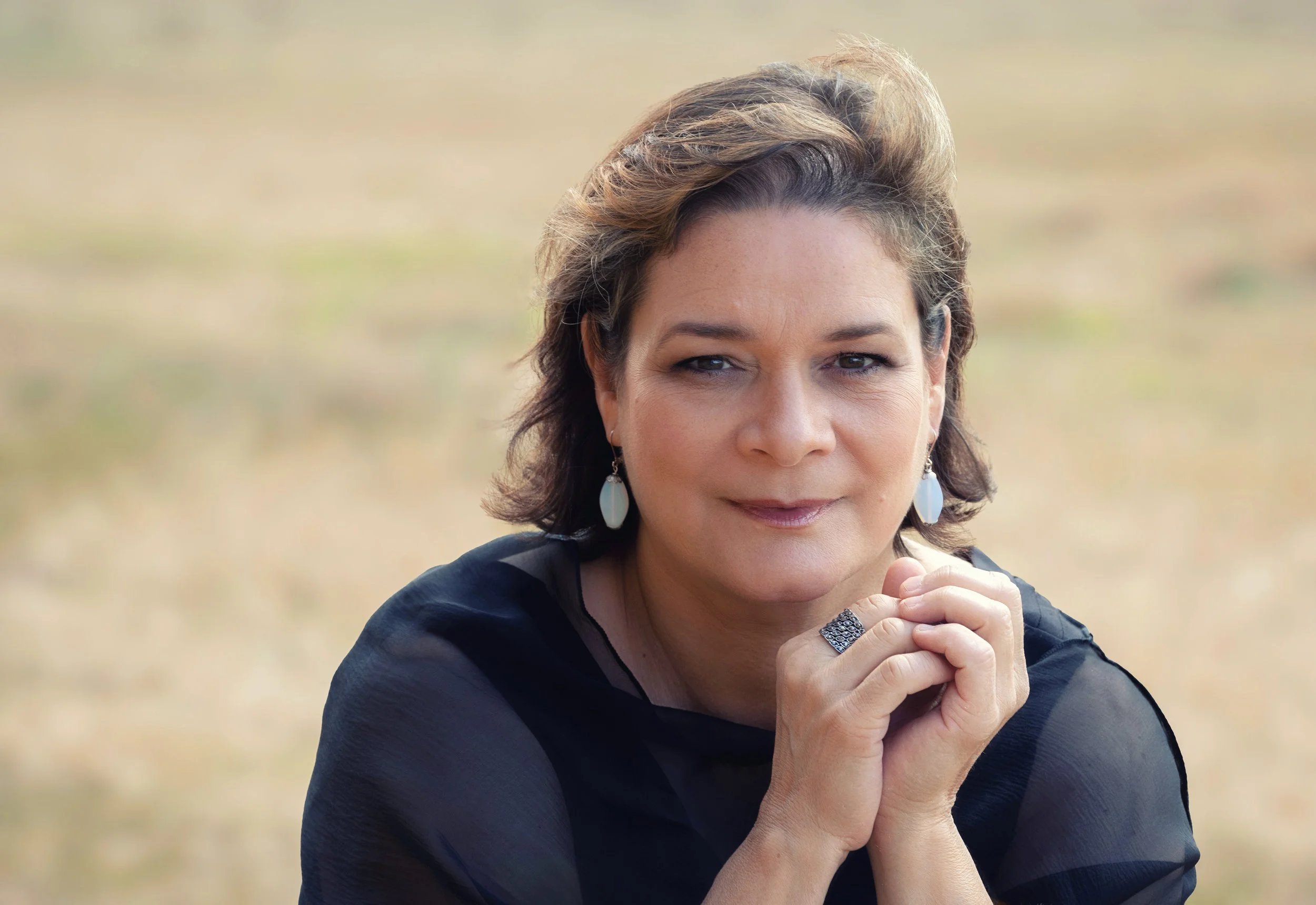 A woman outdoors with short brown hair, wearing earrings and a ring, resting her chin on her clasped hands, looking at the camera with a slight smile.