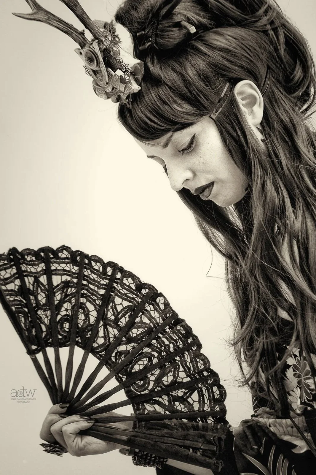 A black and white photo of a woman with long hair wearing glasses and a decorative headpiece, holding a lace fan, with her eyes closed.