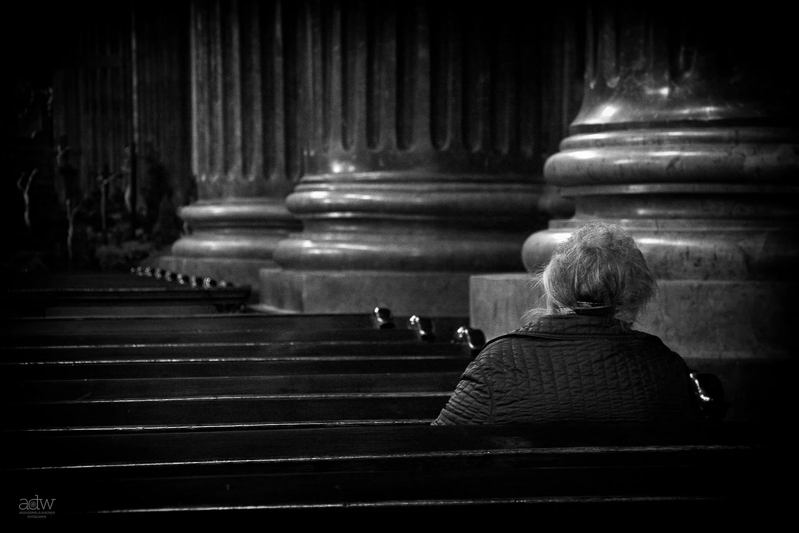 A person with gray hair and a dark quilted jacket sitting on a church pew inside a dimly lit cathedral, with large columns in the background.