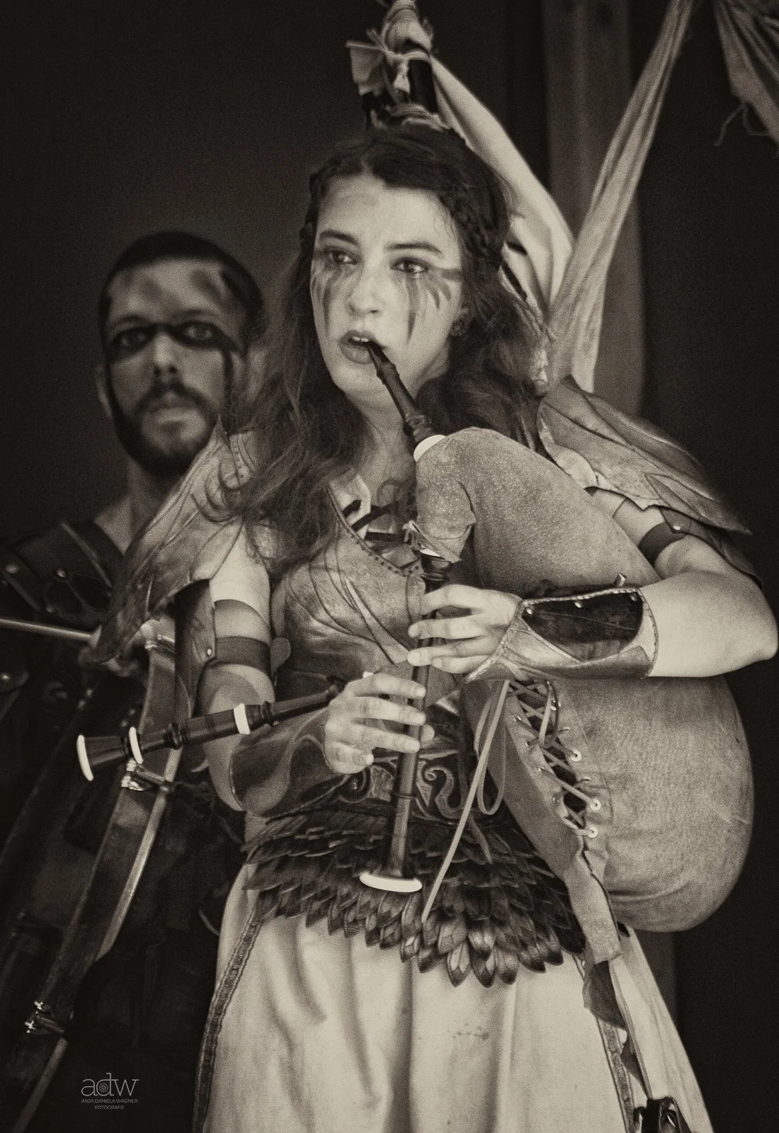 A woman dressed in fantasy warrior costume with feathered skirt and arm guards, holding a sword and smoking a pipe, standing in front of a man in armor with glasses, both with painted faces.