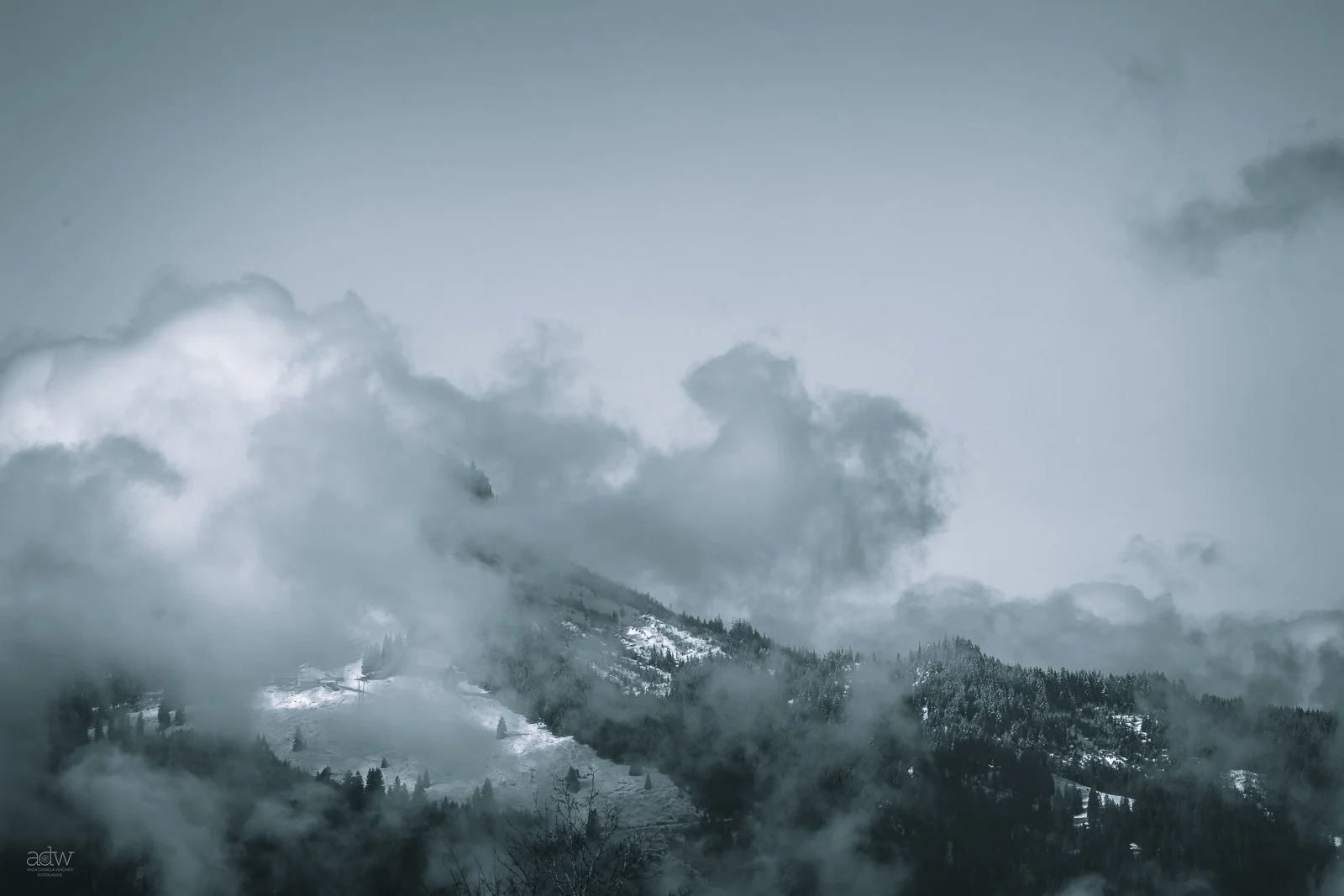 Mountain landscape with snow-covered peaks, trees, and thick clouds or fog.