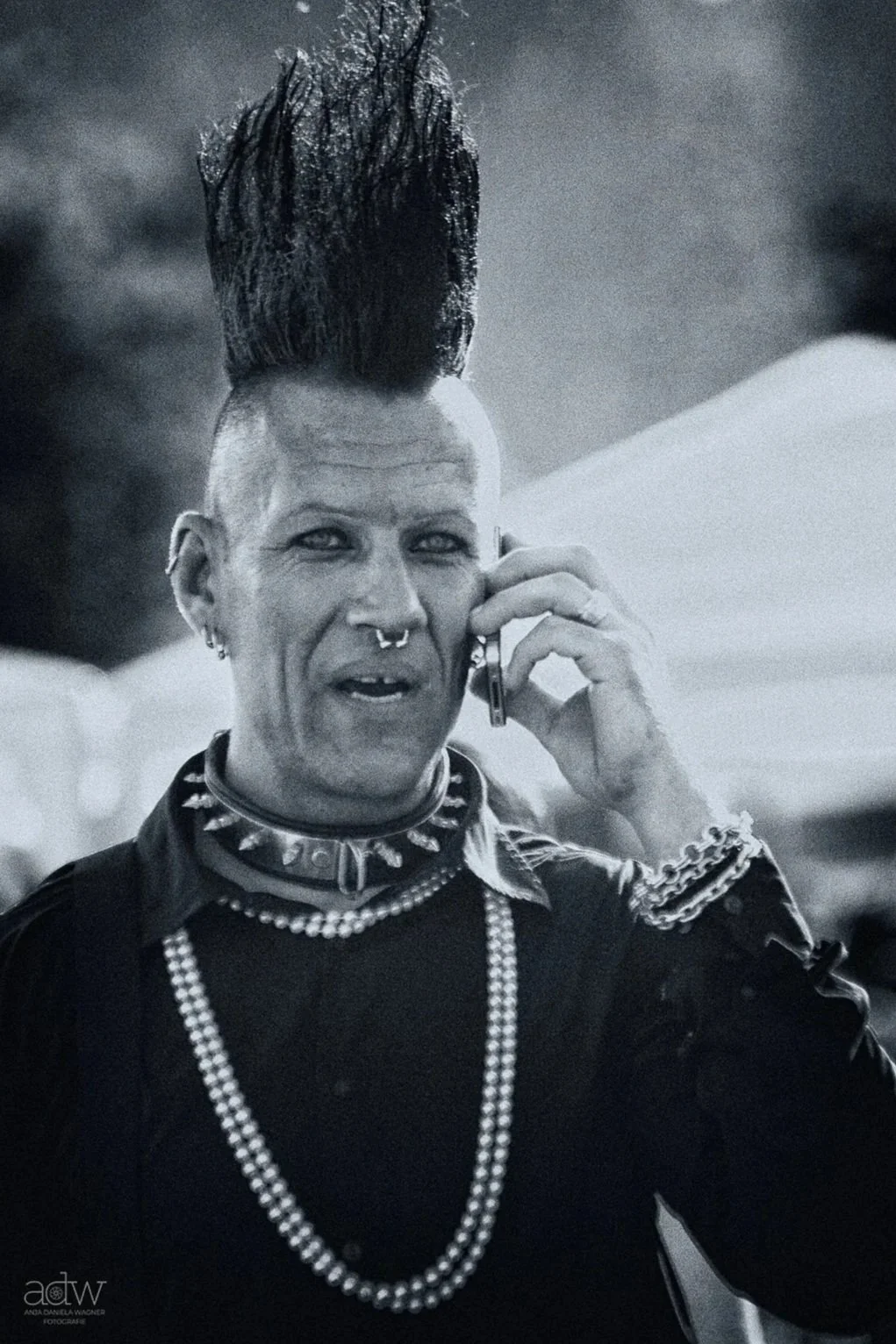 A person with a mohawk hairstyle, piercings, and many necklaces is talking on a cell phone.