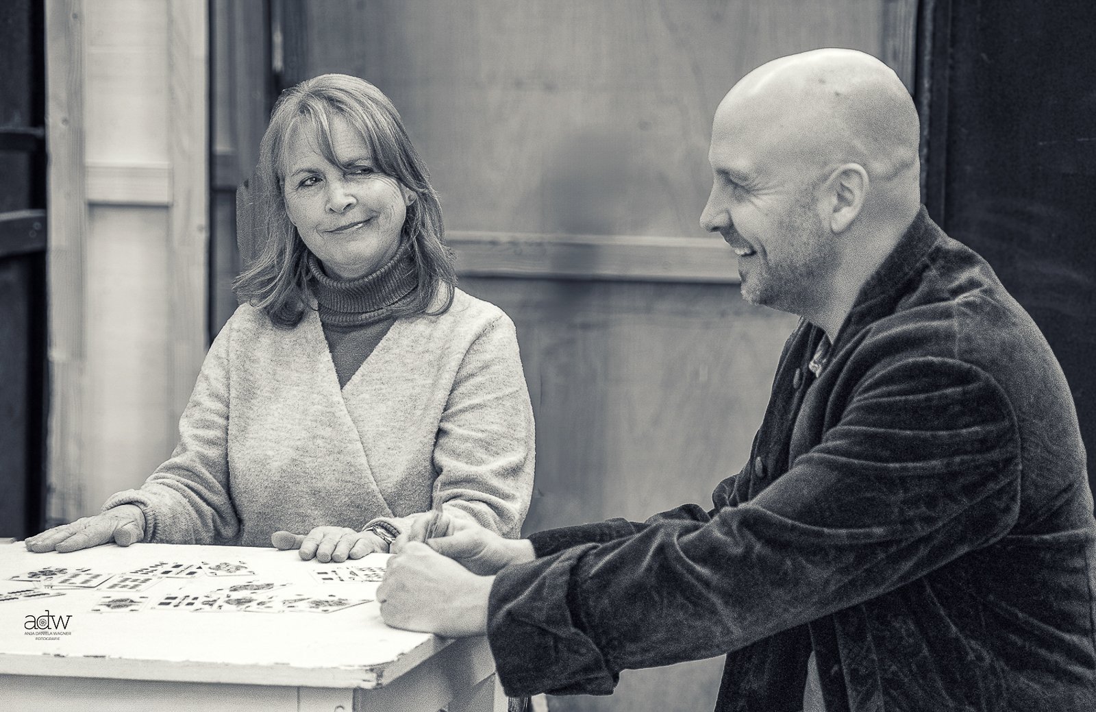 A black and white photo of a woman and a man sitting at a table. The woman is winking and smiling, wearing a turtleneck sweater and a blazer. The man has a bald head, is smiling, and is wearing a jacket. They appear to be playing a game, possibly Scrabble, on the table.