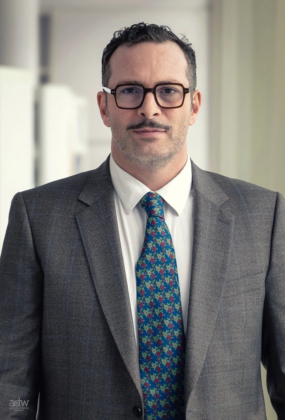A man with short dark hair, glasses, and facial hair, wearing a gray blazer with a checkered pattern, a white shirt, and a colorful floral tie, standing indoors with a blurred background.