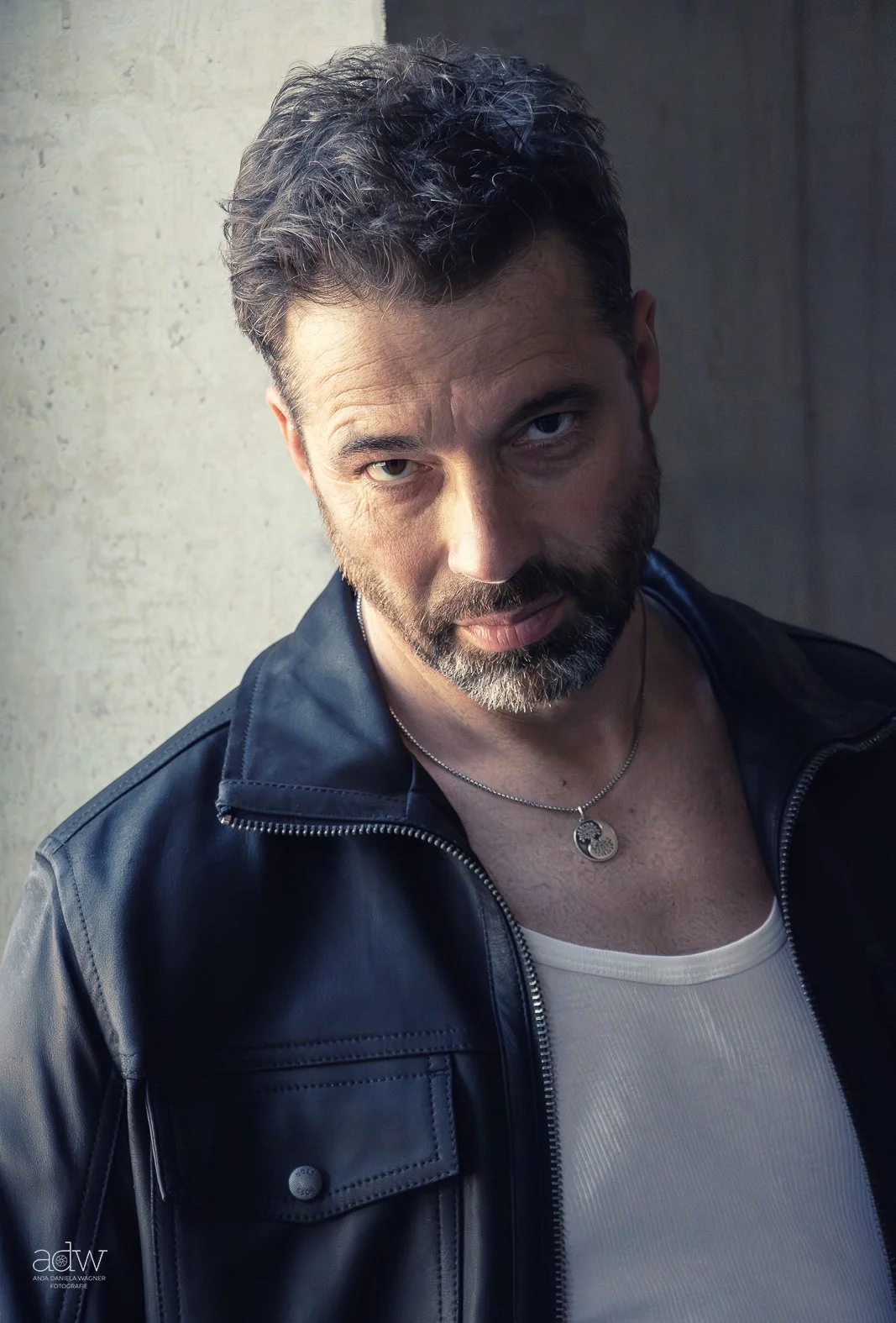 A man with dark hair and a beard wearing a black leather jacket, white T-shirt, and a necklace with a circular pendant posing against a wall.