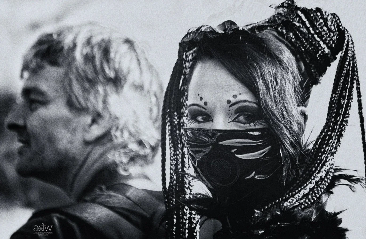 Black and white photo of two people, with one person wearing a mask and featuring elaborate braided hair and face makeup, and the other person with dreadlocks facing sideways.