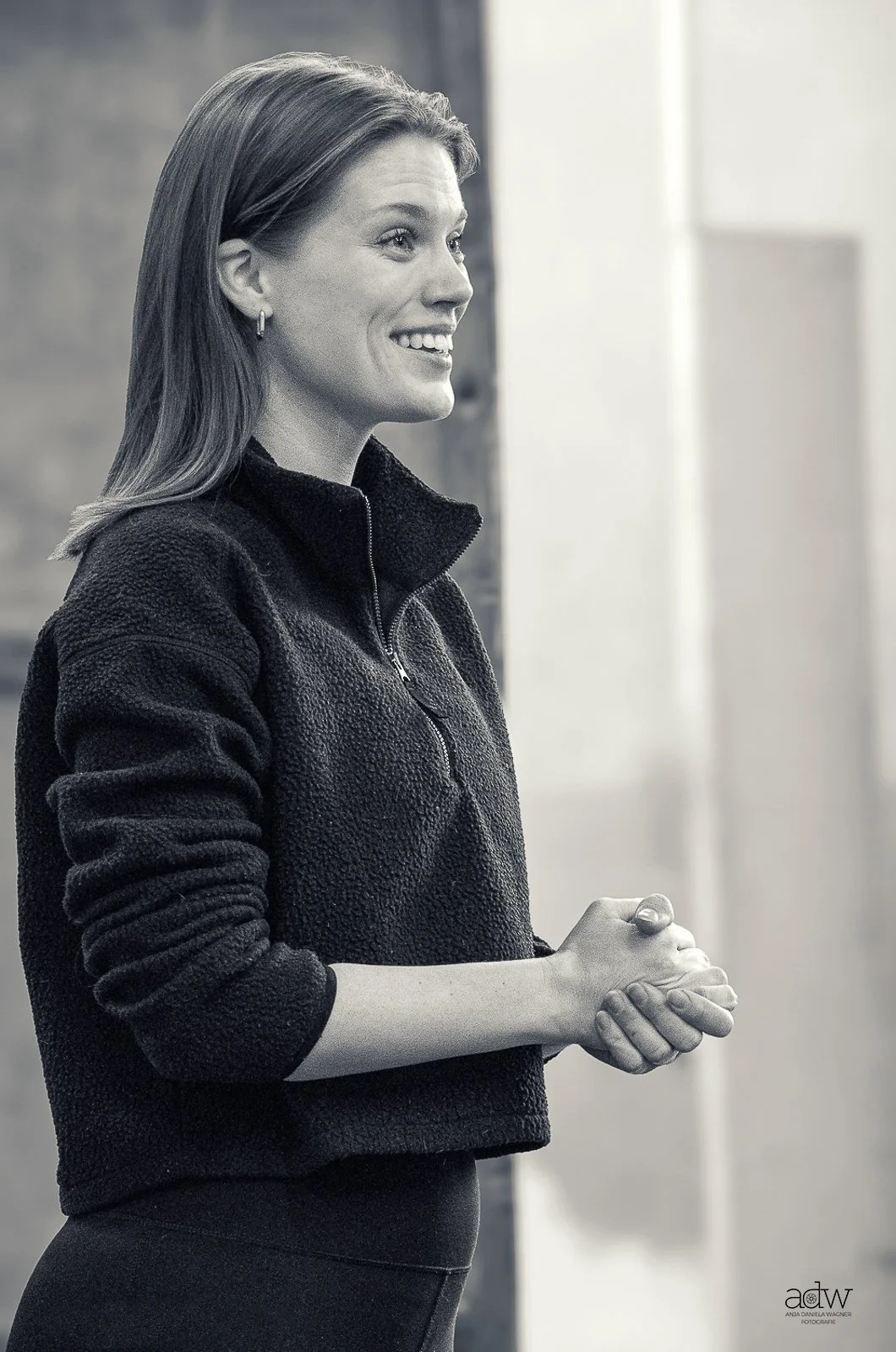 A woman with shoulder-length hair, wearing a zippered fleece jacket, is smiling and clasping her hands together.