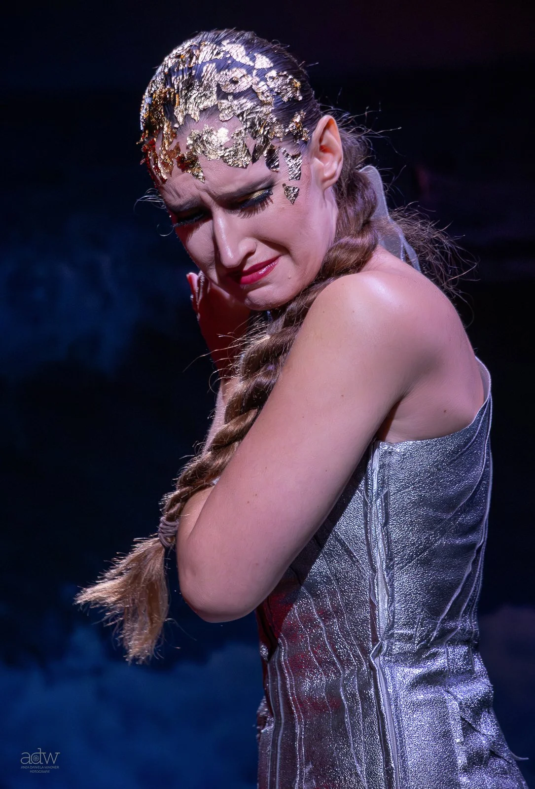A woman with gold leaf on her forehead, wearing a shimmering silver dress, with her eyes closed and a contemplative expression.
