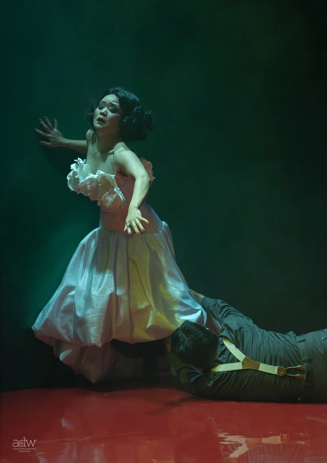 A woman in a vintage-style pink dress is kneeling on the floor with a man lying underneath her. The woman appears distressed, with one hand on her stomach and the other outstretched. The man is partially visible, lying face down on the ground, dressed in dark clothing with a yellow strap.
