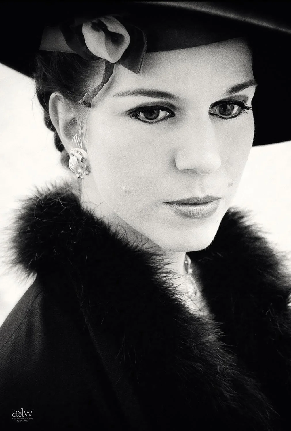 Black and white portrait of a woman with vintage style, wearing a wide-brimmed hat with a bow, earrings, and a fur collar.