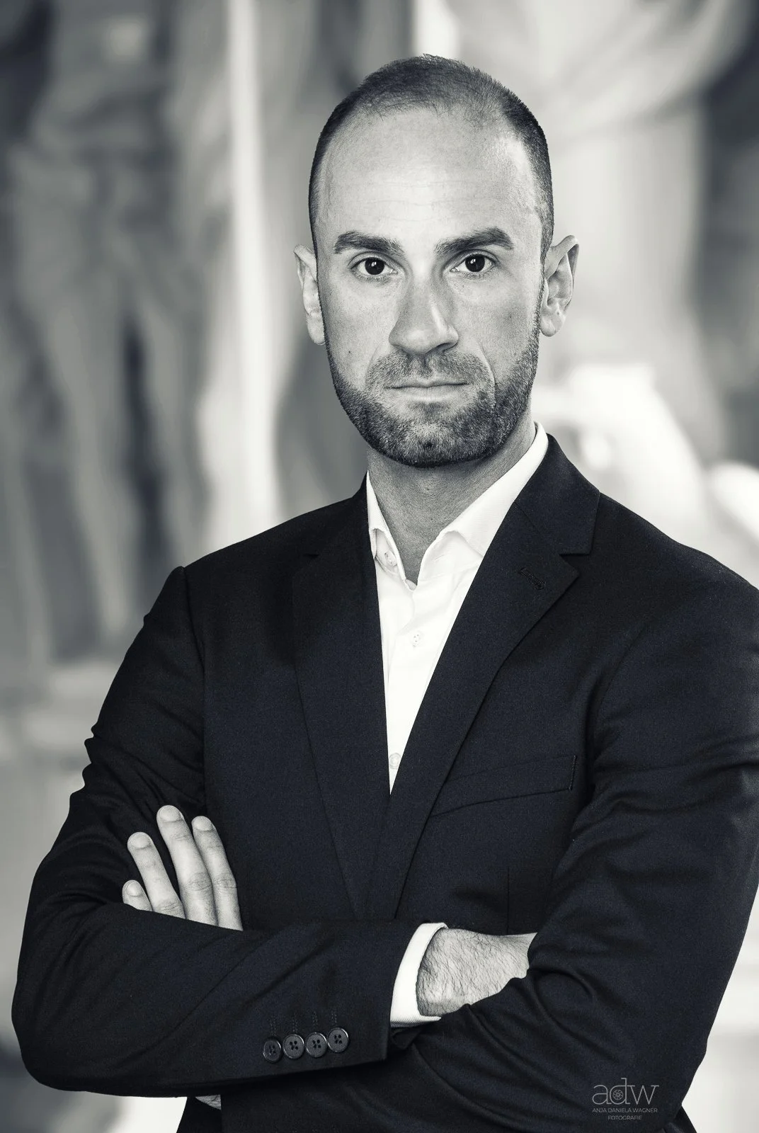 Black and white portrait of a man with short hair and a beard, wearing a suit with arms crossed, looking directly at the camera.